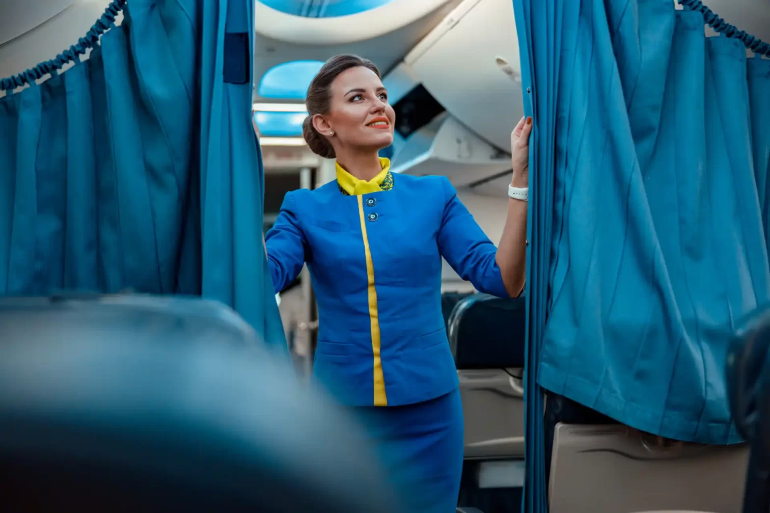 Comissária de bordo feminina sorridente posando dentro de uma aeronave, destacando-se por seu uniforme azul com detalhes amarelos, simbolizando hospitalidade e segurança.