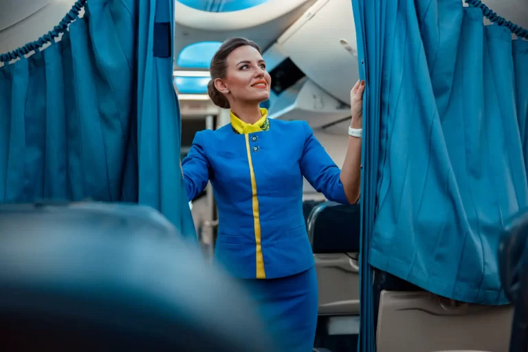 Comissária de bordo feminina sorridente posando dentro de uma aeronave, destacando-se por seu uniforme azul com detalhes amarelos, simbolizando hospitalidade e segurança.