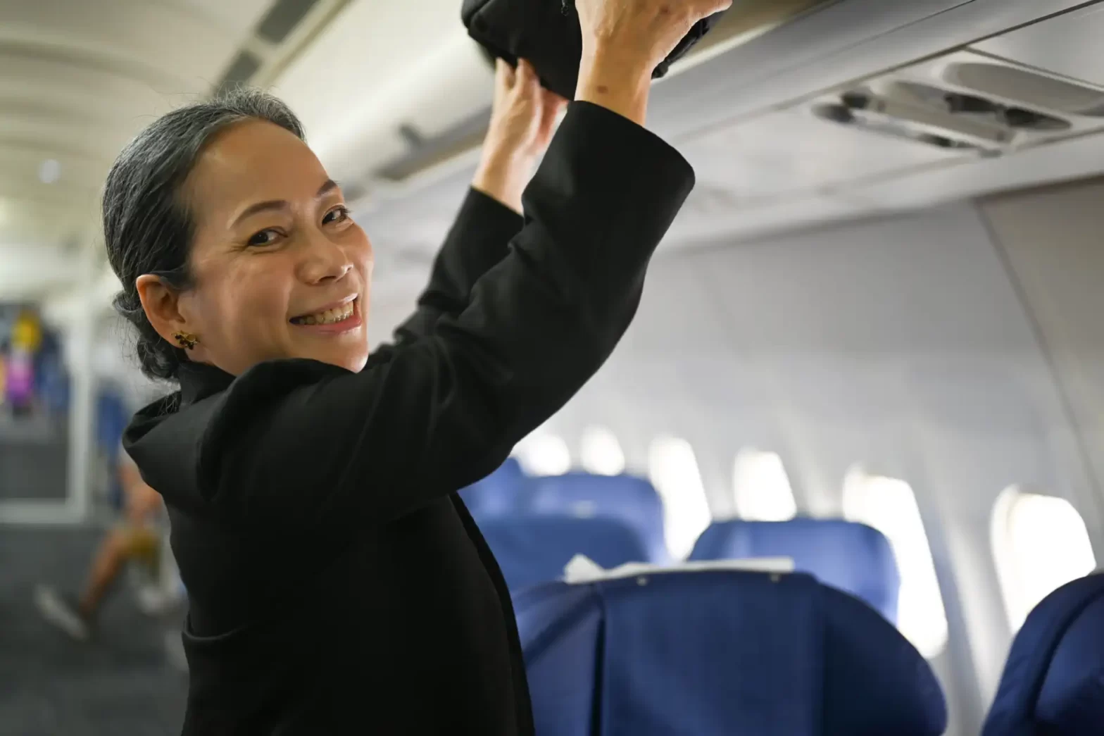 Comissária de bordo sorridente organizando bagagens no compartimento de um avião, transmitindo hospitalidade e eficiência aos passageiros durante o voo.