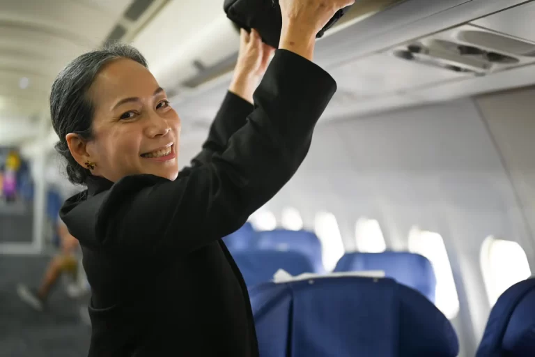 Comissária de bordo sorridente organizando bagagens no compartimento de um avião, transmitindo hospitalidade e eficiência aos passageiros durante o voo.