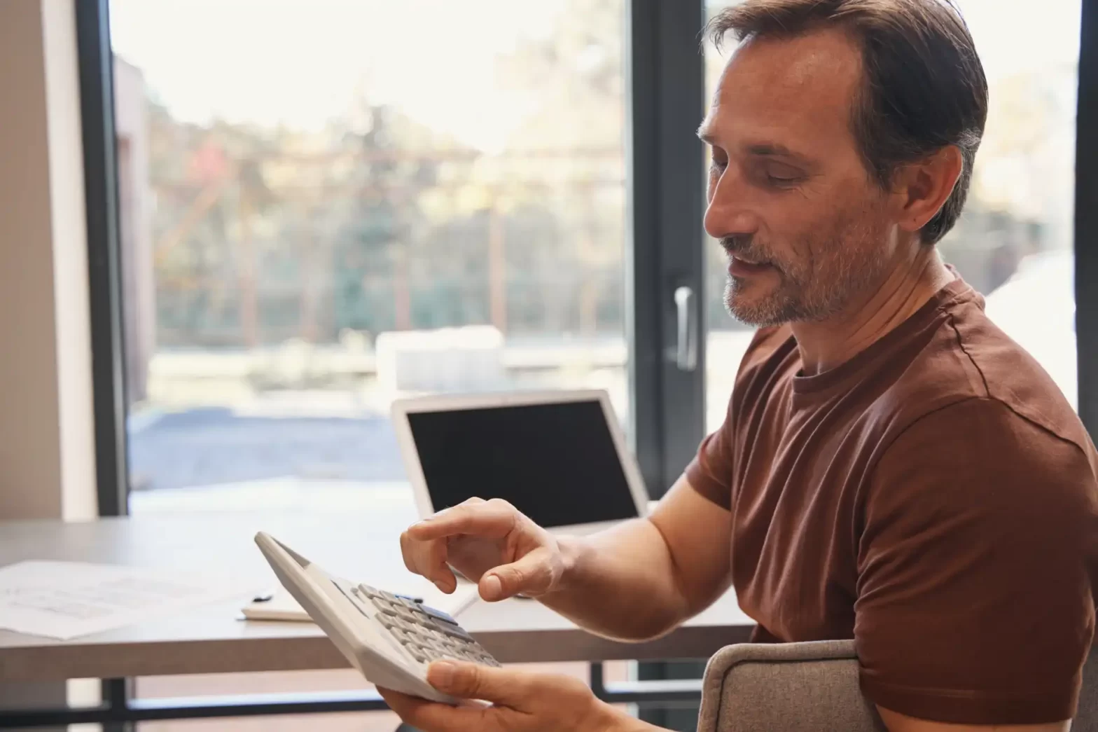 Homem de meia-idade usando calculadora em escritório com grande janela ao fundo, ambiente iluminado e moderno, focado em cálculos e finanças.
