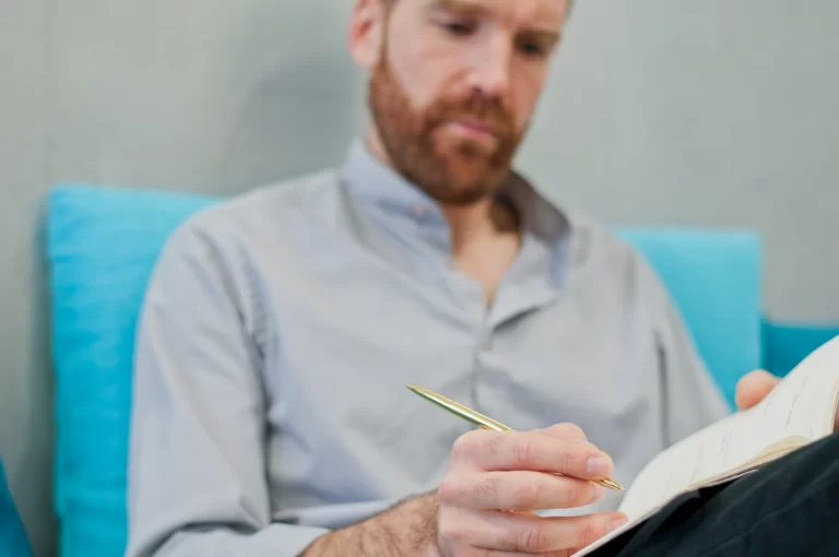 Homem concentrado escrevendo em um caderno, sentado em um sofá. Ele usa uma camisa clara e segura uma caneta. O ambiente é moderno e acolhedor.