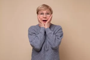 Mulher idosa mostrando expressão de surpresa com as mãos no rosto, vestindo um suéter cinza em fundo neutro, simbolizando emoções e reações inesperadas.