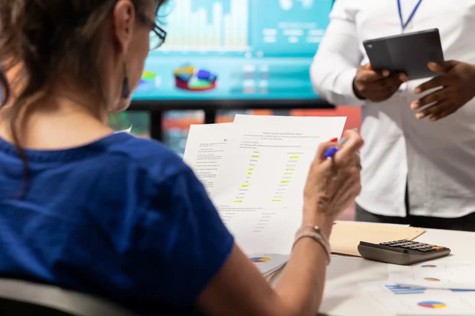 Pessoa analisando relatórios financeiros em uma reunião de negócios, com gráficos no computador ao fundo, focada em gestão financeira e análise de dados.