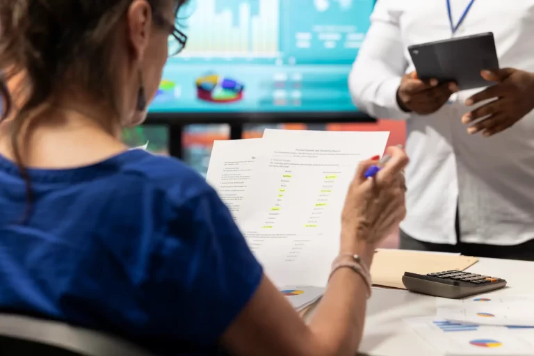 Pessoa analisando relatórios financeiros em uma reunião de negócios, com gráficos no computador ao fundo, focada em gestão financeira e análise de dados.