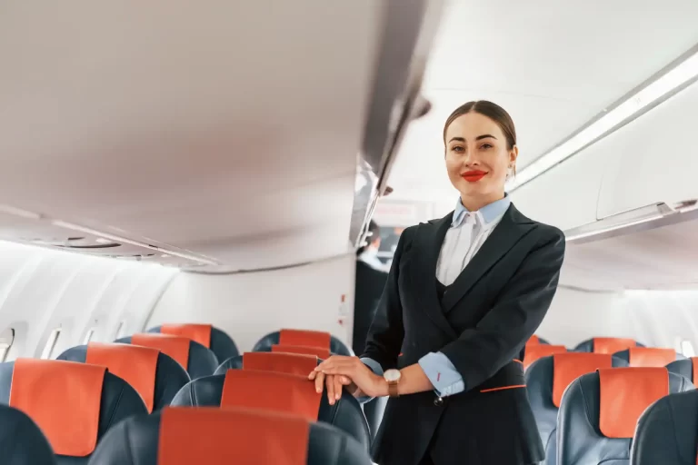 Comissária de bordo sorridente dentro de uma aeronave, destacando-se pelo uniforme formal e ambiente de avião com assentos azuis e detalhes em laranja.