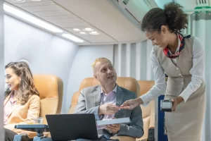 Executiva de cabine de avião atendendo passageiro durante voo, com outros passageiros ao fundo, ambiente de avião moderno e confortável. Aposentadoria do comissário de bordo por Insalubridade.