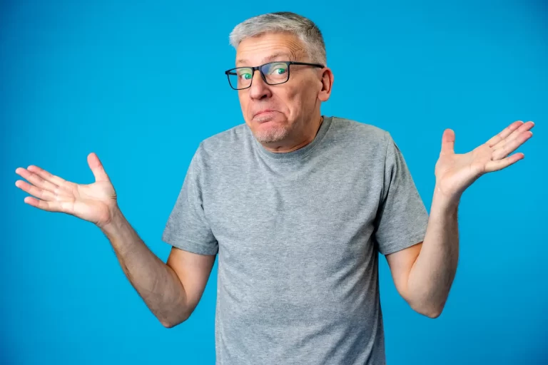 Homem com expressão de dúvida, vestindo camiseta cinza e óculos, com cabelo cinza claro, gesticulando com as mãos de forma incerta contra fundo azul. Tipos de Aposentadoria no Brasil.