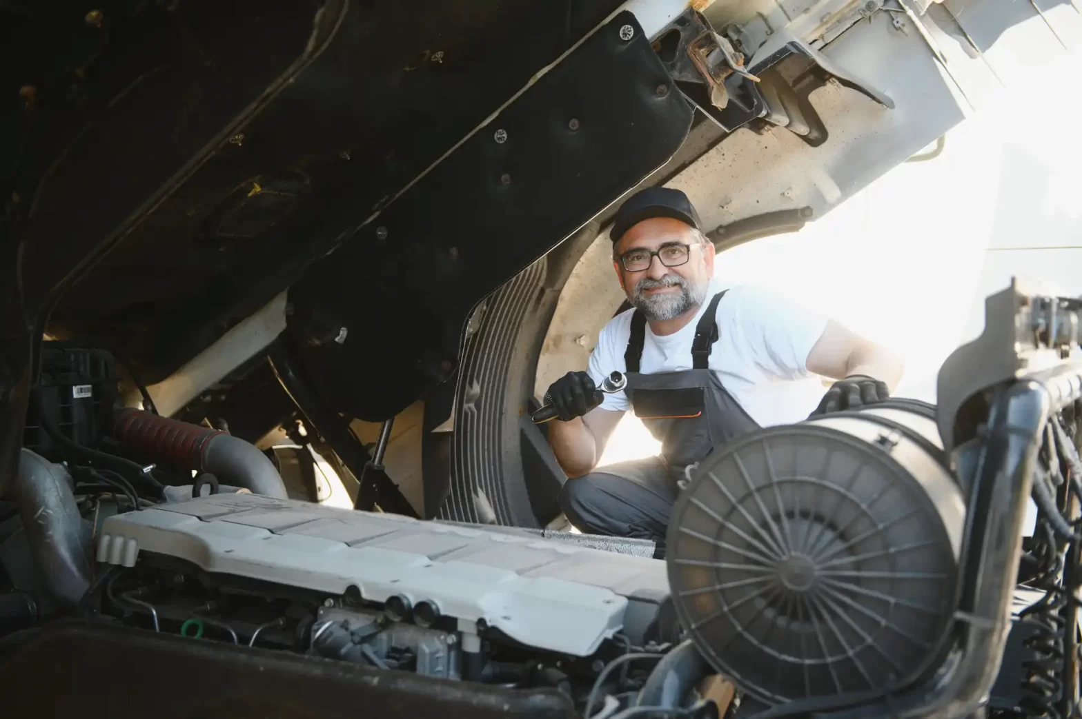 Aposentadoria Especial dos Aeroviários. Homem mecânico com barba e óculos analisando motor de carro sob o capô aberto, usando uniforme de trabalho na oficina mecânica.