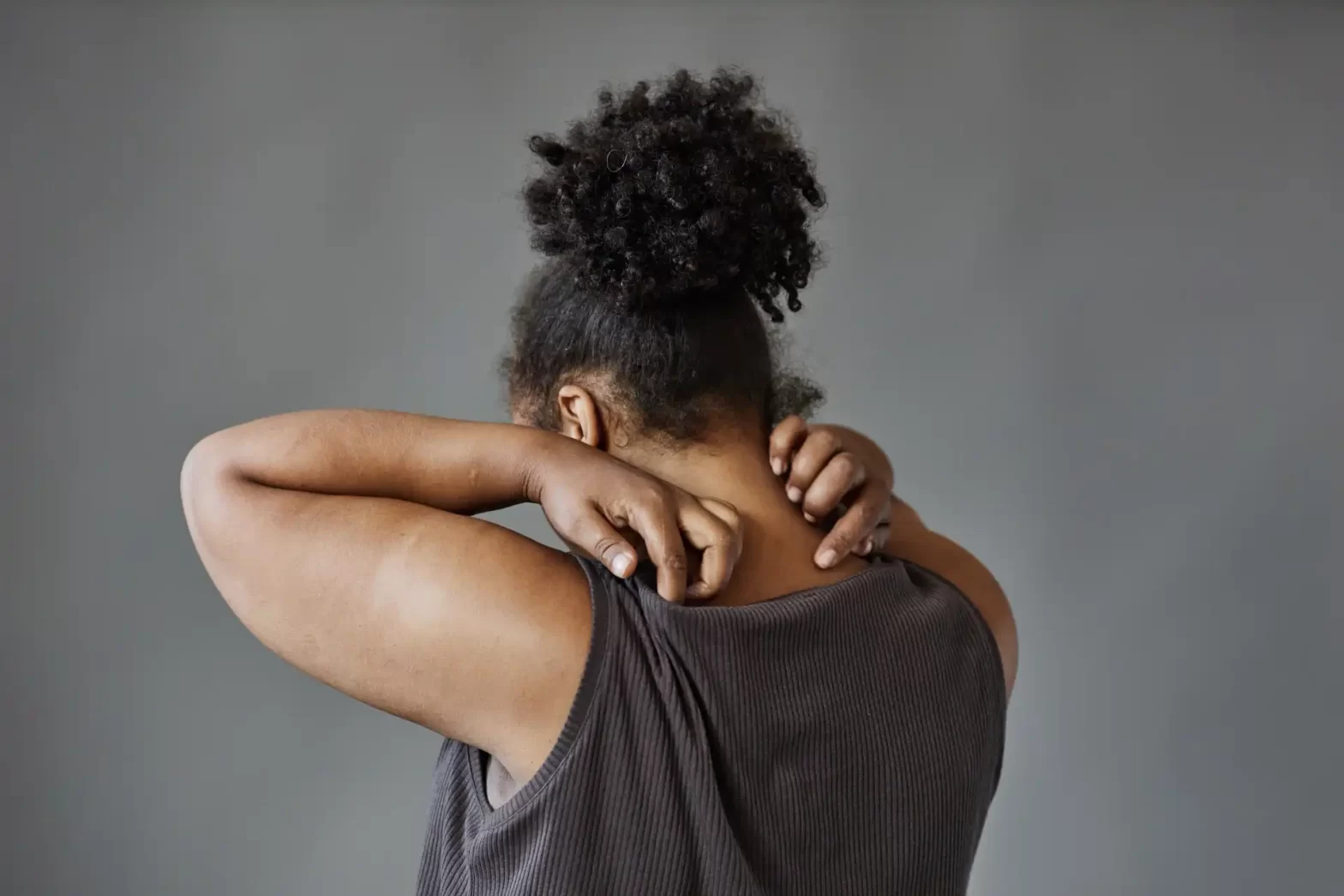 Mulher segurando a região do pescoço e ombro, demonstrando desconforto ou dor, com cabelo afro e vestindo uma blusa cinza escura contra fundo cinza. Post fibromialgia como deficiência.