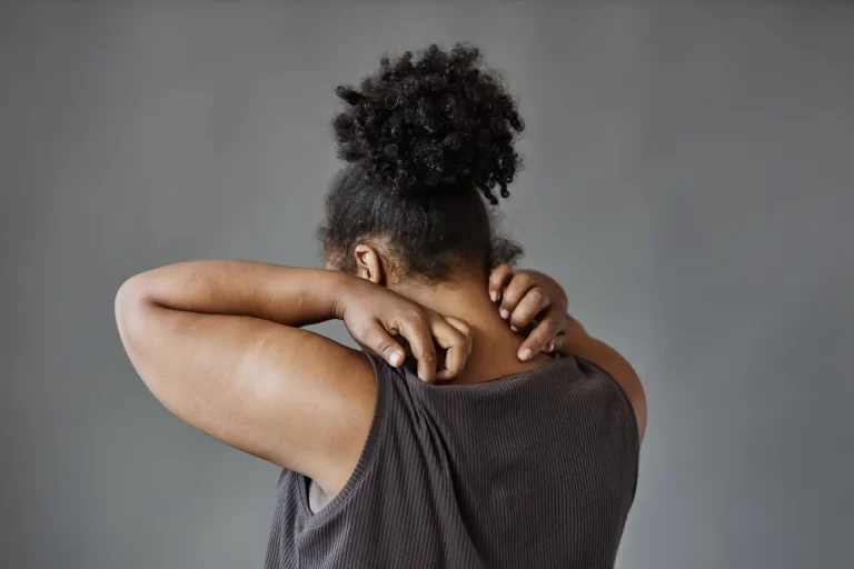 Mulher segurando a região do pescoço e ombro, demonstrando desconforto ou dor, com cabelo afro e vestindo uma blusa cinza escura contra fundo cinza. Post fibromialgia como deficiência.