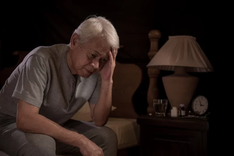 Imagem de uma pessoa idosa com expressão preocupada ou pensativa, sentada na cama ao lado de uma luminária e um relógio de parede, transmitindo um momento de reflexão ou tristeza. Erros ao planejar a aposentadoria.