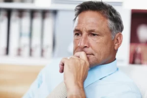 Homem pensativo de camisa azul, demonstrando reflexão e preocupação, sentado em escritório com estantes ao fundo. Inflação e Aposentadoria.