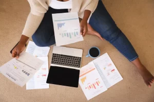 Elisão fiscal x evasão. Pessoa analisando gráficos financeiros e relatórios de negócios com laptop e xícara de café, em ambiente de trabalho doméstico.