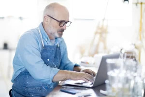 Homem idoso trabalhando em um computador portátil na sua oficina ou ateliê, usando avental e óculos, com materiais de arte ao fundo. Post.: Planejamento Previdenciário para quem começou a trabalhar tarde