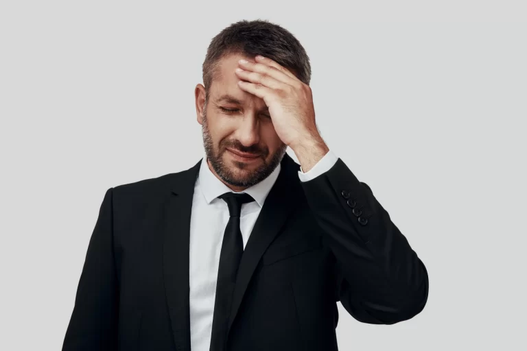 Homem de terno preto com rosto de dor ou preocupação, segurando a cabeça com a mão, expressando frustração ou estresse.