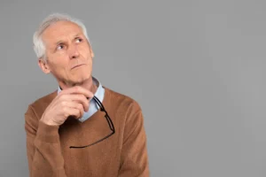 Homem idoso com cabelo grisalho, em estúdio, usando suéter marrom e camisa azul, segurando um pequeno fone de segurança com microfone na mão, com expressão pensativa e fundo cinza liso.