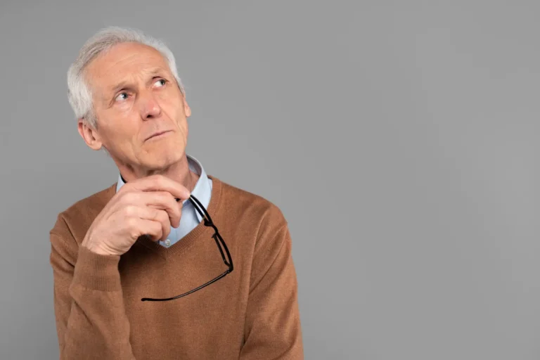 Homem idoso com cabelo grisalho, em estúdio, usando suéter marrom e camisa azul, segurando um pequeno fone de segurança com microfone na mão, com expressão pensativa e fundo cinza liso.