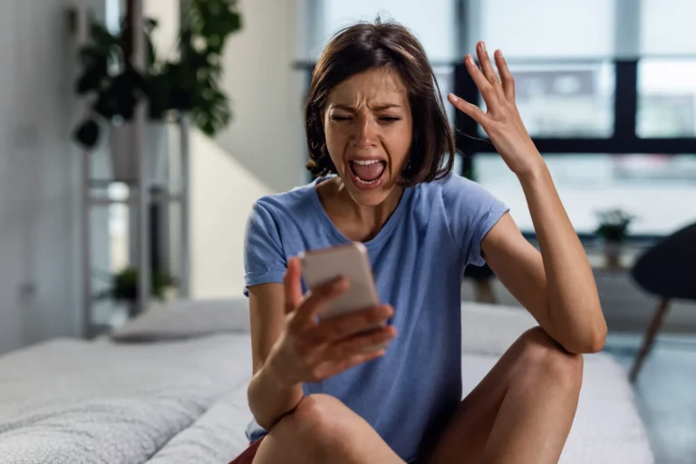 Mulher sentada na cama em um quarto moderno, com expressão de dor e assustada, usando o celular na mão enquanto tenta se afastar ou tapar o ouvido com a outra mão, sugerindo estresse ou desconforto