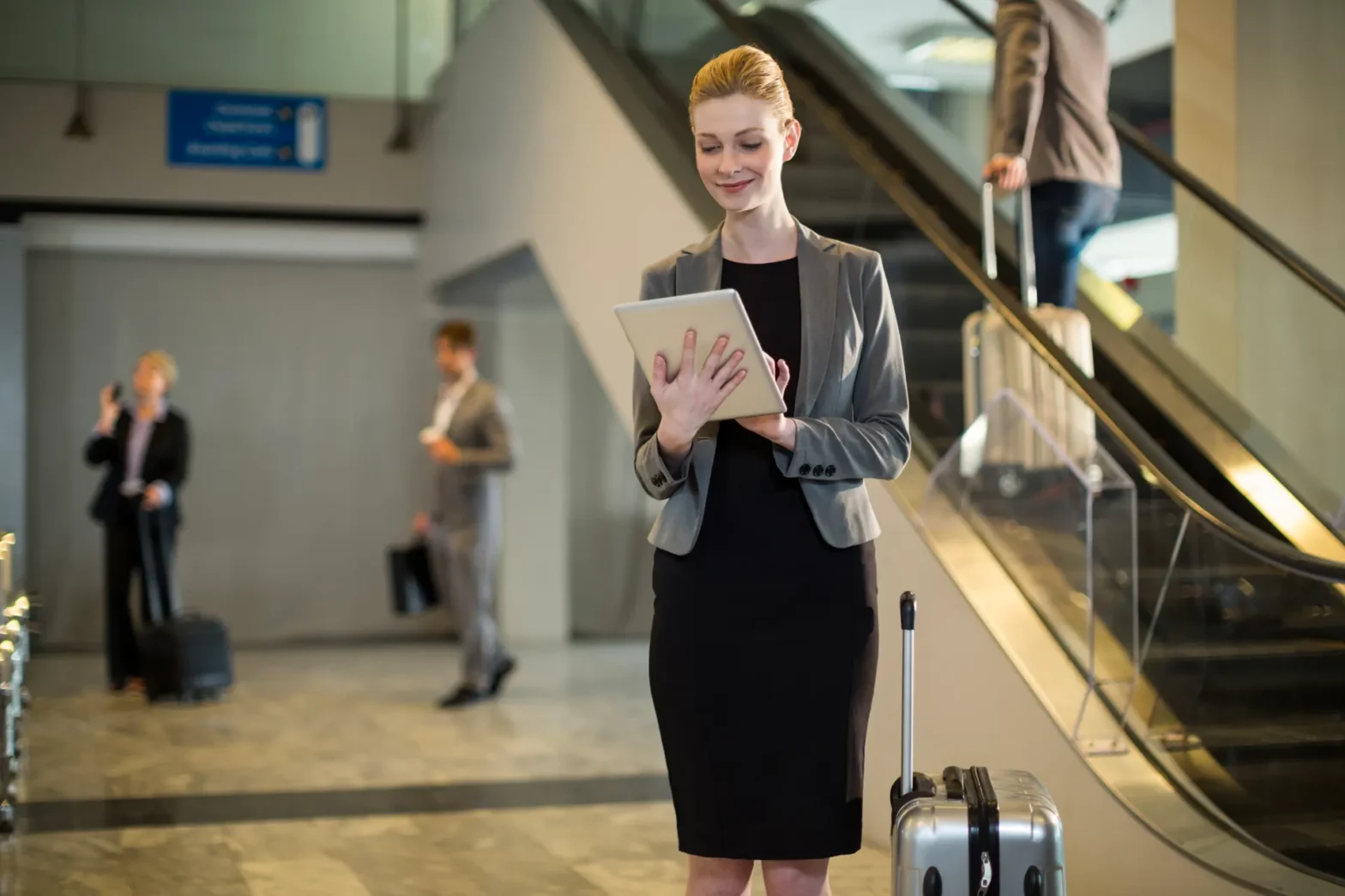 Mulher executiva com terno e vestido preto caminha em aeroporto, segura uma pasta e sorri enquanto puxa uma mala de viagem ao lado de escadas rolantes. Blogpost "Tempo em solo".