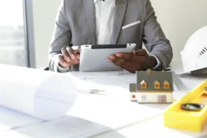 Engenheiro ou arquiteto em reunião usando tablet sobre plantas arquitetônicas, enquanto equipamentos e um modelo de casas em miniatura estão sobre a mesa, ao lado de capacete branco e ferramentas. Planejamento Sucessório.
